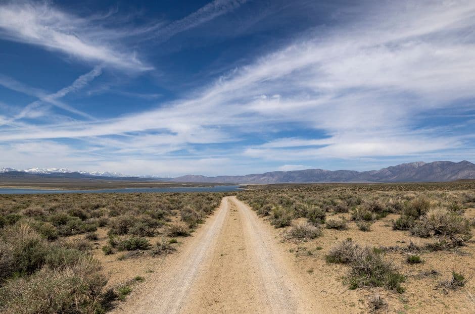 A dirt road stretches through the desert landscape towards distant snowy mountains under a clear sky.